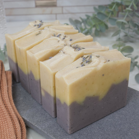 Five Soap bars are stacked one in front of the other on a grey stone surface. The soaps feature a layered pattern with dark purple base and bright yellow top. Lavender pieces can be seen on top of the soaps