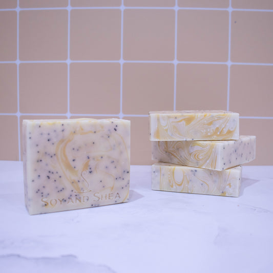 Three soap bars lay stacked upon each other showing a flat while top with yellow swirl. To the left stands a fourth bar which is white with yellow wisps of colour and black flecks of poppy seeds. These sit on a marble surface with pale orange tile background.