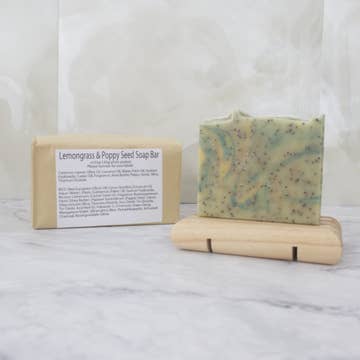 A pale green soap bar with green and yellow swirls and black flecks sits on a wooden stand with a brown paper package behind labeled 'Lemongrass and Poppy Seed Soap Bar' on a marble background.