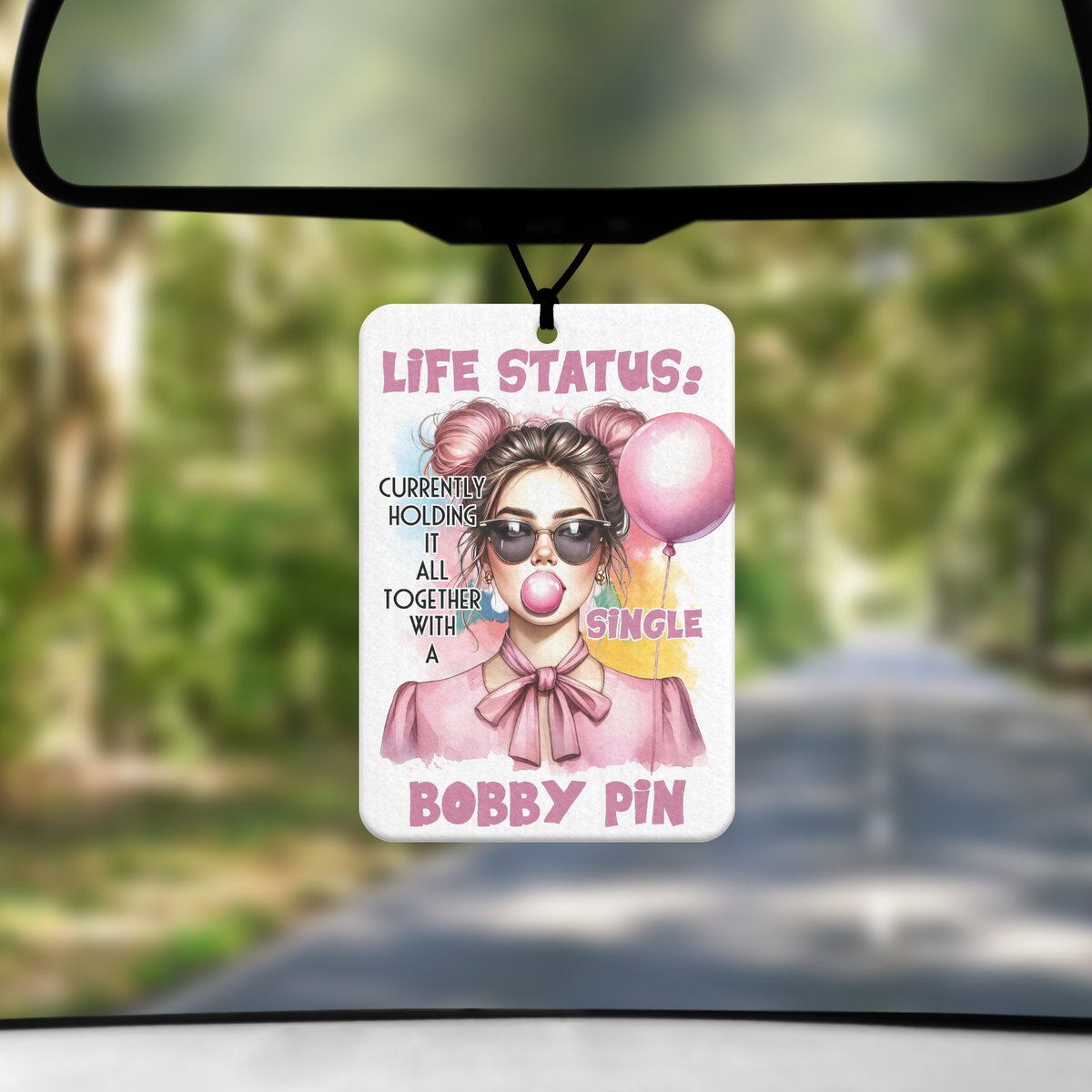 A mock up image showing a view through a car windscreen with a long tree lined road. From the rearview mirror hangs an air freshener with an image ofof a youn lady blowing on bubblegum and holding a pink balloon. The words LIFE STATUS is across the top followed by "currently holding it all together with a single Bobby pin"