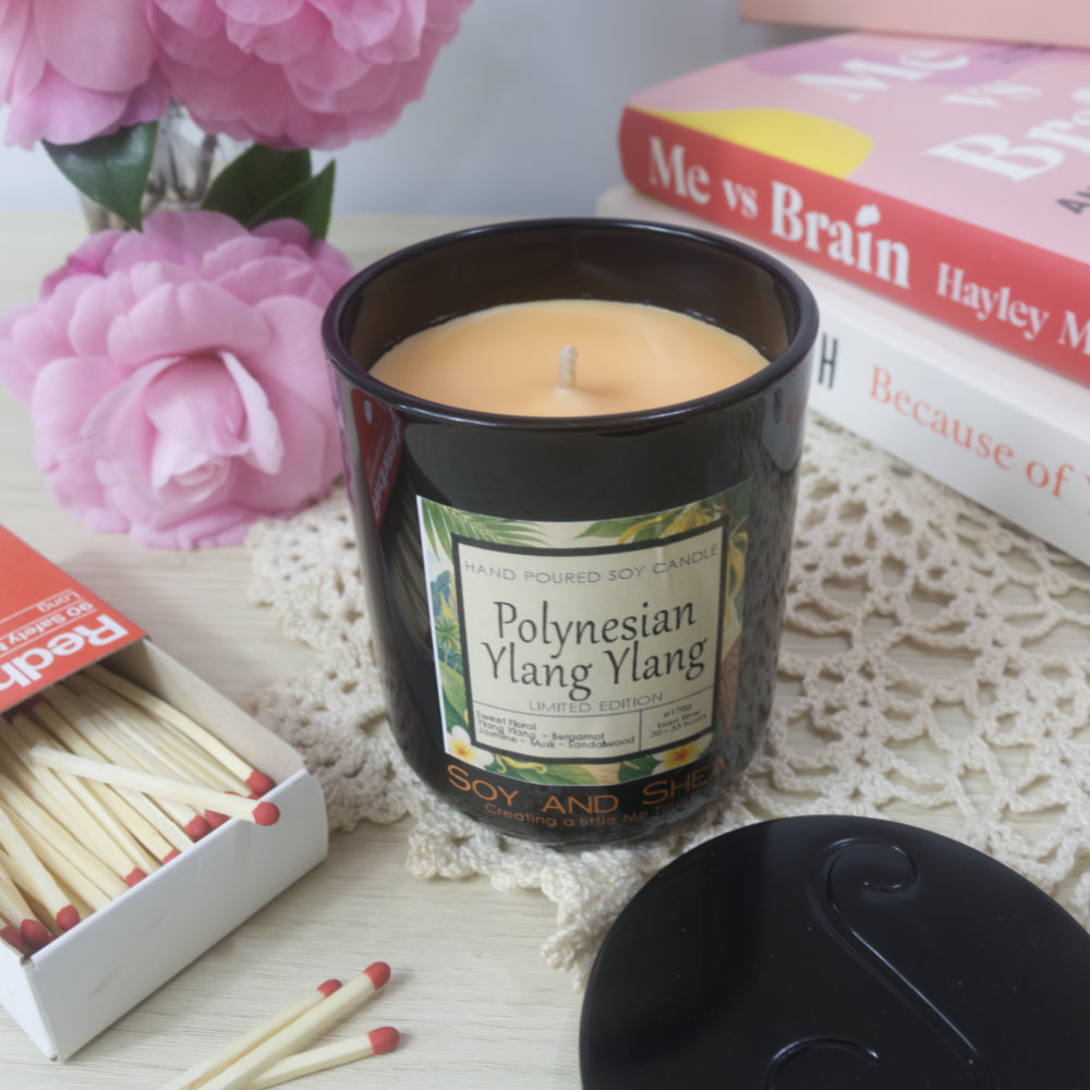 A black candle jar with decorative label is on a sideboard with vase of pink flowers to the left and a stack of bookes to the right. The lid of the jar has been removed and placed to the right in front of the books revealing a light orange wax.