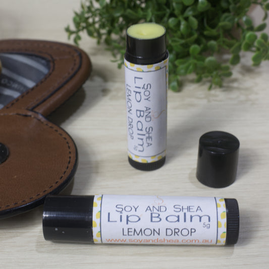 Two black lip balm tubes labeled 'Soy and Shea Lip Balm Lemon Drop' on a wooden surface with a brown leather bag and green plant in the background. One Lip Balm is lying on its side while the other is standing on its end with the lid removed showing a yellow balm.