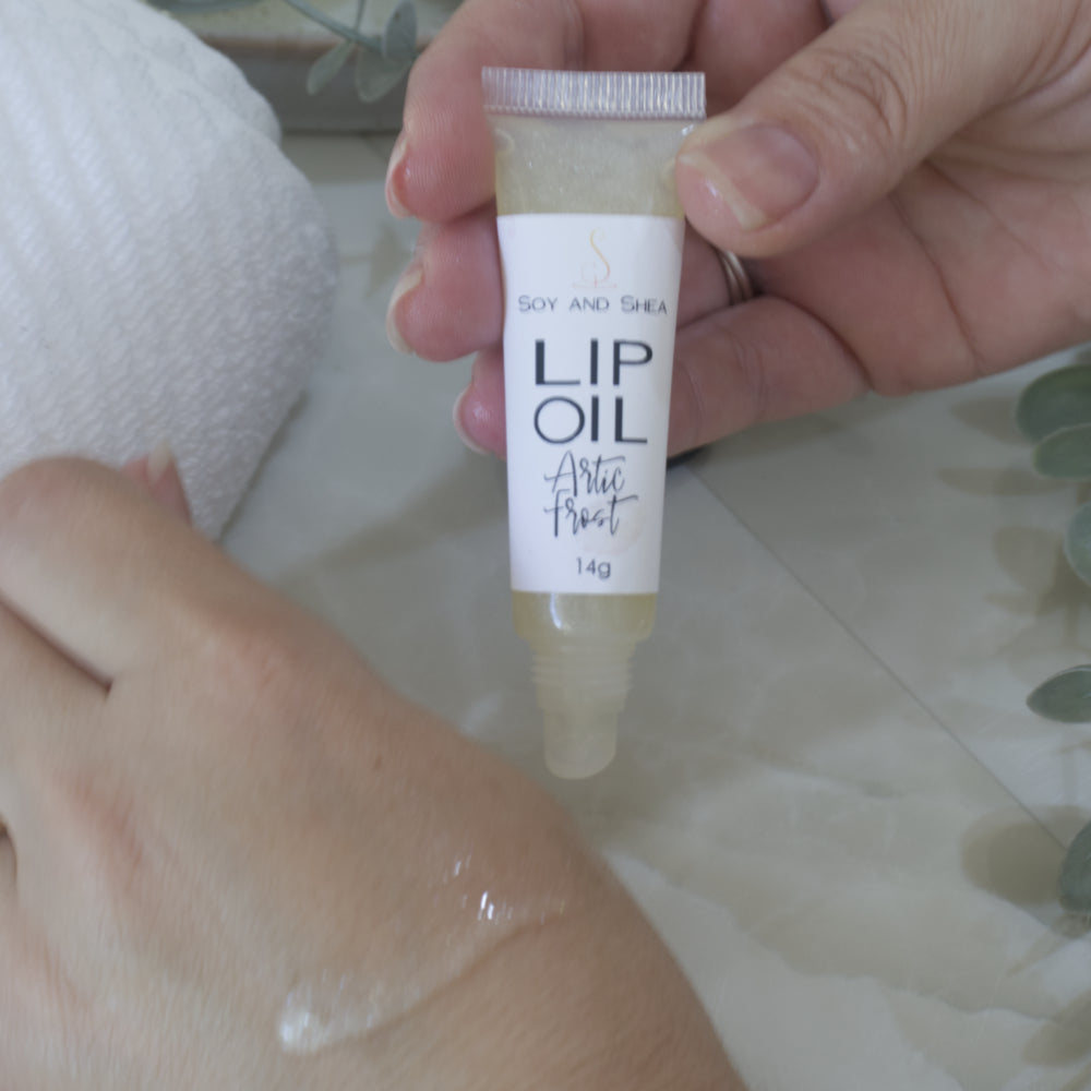A hand holds a tube of Lip oil next to another hand that has a smear of the oil across the back demonstrating the colour and translucent nature of the oil. The tube is clear showing an off-white oil that goes onto the hand amost clear with a sparkle