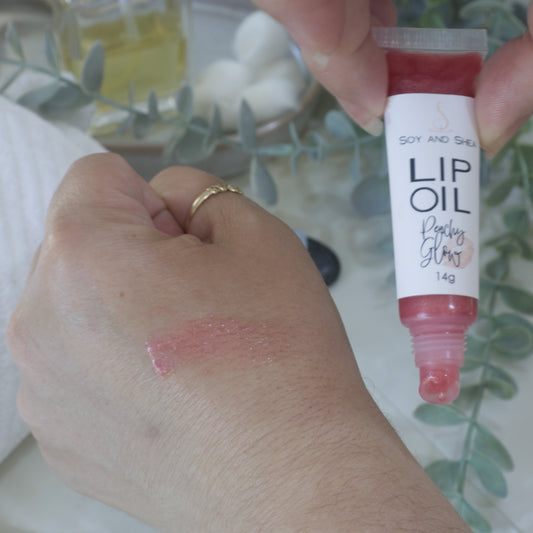 A hand holds a tube of Lip oil next to another hand that has a smear of the oil across the back demonstrating the colour and translucent nature of the oil. The tube is clear showing a peachy red Lip Oil that goes onto the hand with a sheer look.