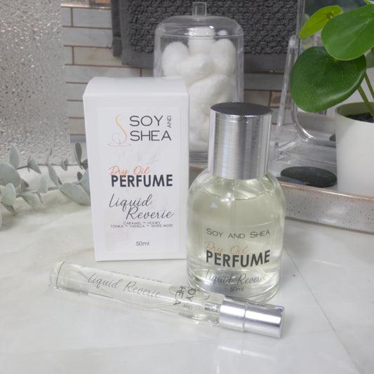 A selection of perfume bottles are presented on a bathroom countertop. To the front is a clear, narrow, tall bottle labeled as 10ml with a silver cap and behind is a clear round, short bottle with silver lid labelled as 50ml. Behind sits a white box with label identifying as Dry Oil Perfume Liquid Reverie.