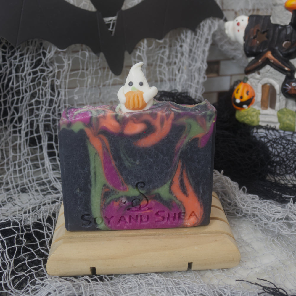 A bar of stand stands on a wooden soap rack with a blurred halloween themed background. The soap is black with swirls of neon orange, green and purple. On top of the soap in the centre is a white ghost embellishment that is holding a pumpkin