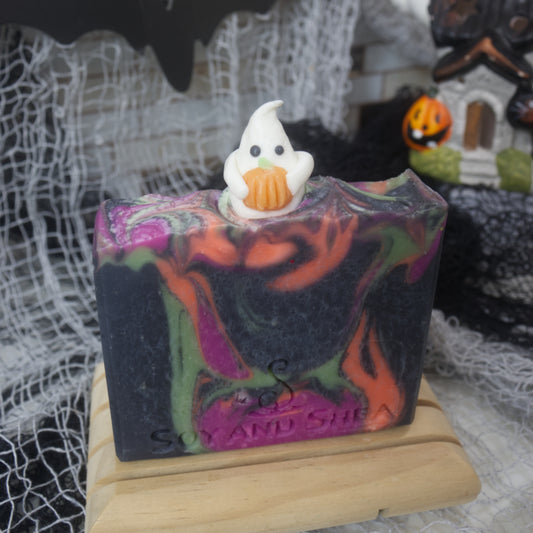 A bar of stand stands on a wooden soap rack with a blurred halloween themed background. The soap is black with swirls of neon orange, green and purple. On top of the soap in the centre is a white ghost embellishment that is holding a pumpkin