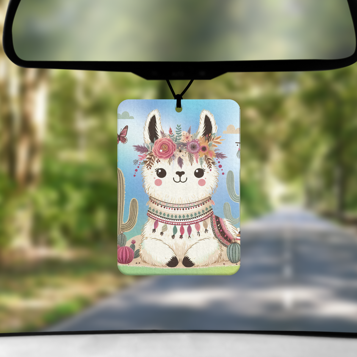 A mock up image showing a view through a car windscreen with a long tree lined road. From the rearview mirror hangs an air freshener with an image of a boho style llama. The llama is white and resting on the ground with a floral headdress and surrounded by cactus