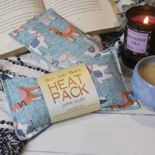 Two Mini Heat packs are stacked upon each other, one with a belly band label reading HEAT PACK LUPIN FIlled. These are resting on a black and white blanket with open book, candle and coffee cup evoking a cosy feel. The Heat packs are pale blue with teal speckles with illustrations of orangle, white and grey llamas wearing blankets their backs