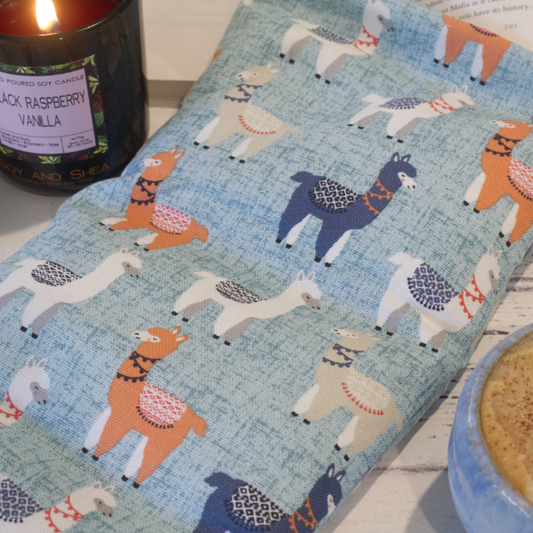 A close up picture of the Llama Drama Heat pack sitting next to a burning candle. The Heat Pack has a light blue speckled background and features drawings of llamas in blue, orange, white and grey