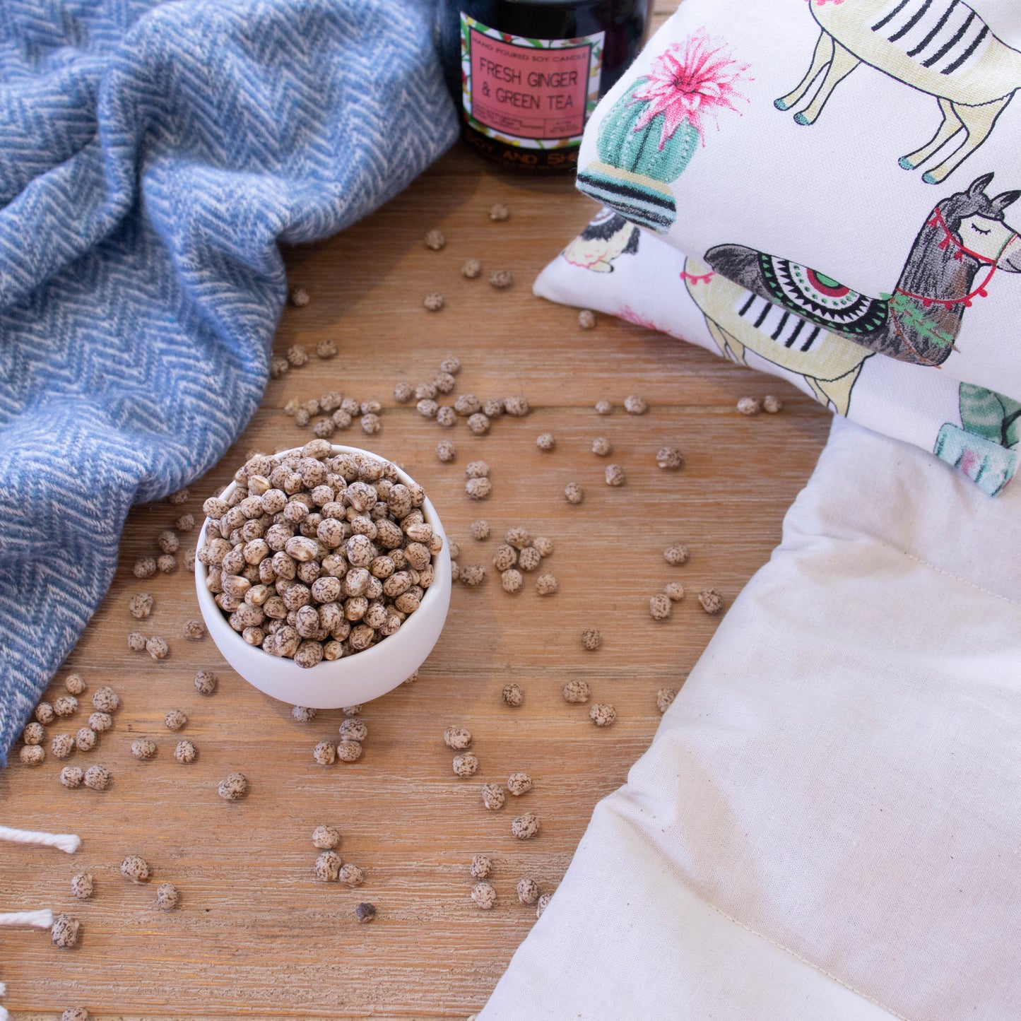 A small white bowl is filled to over flowing with lupin, a small pea sized legume that is tan with brown speckles. The lupin is scattered across a wooden surafce and uis surrounded by a blue blanket and a Heat Pack.