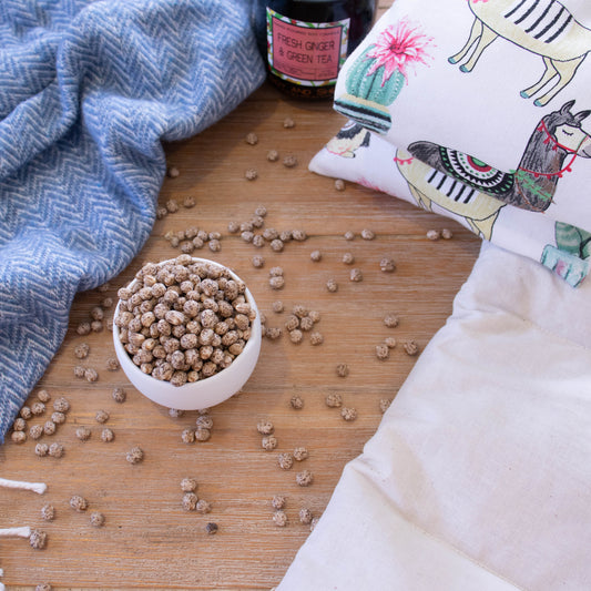 A small white bowl is filled to over flowing with lupin, a small pea sized legume that is tan with brown speckles. The lupin is scattered across a wooden surafce and uis surrounded by a blue blanket and a Heat Pack.