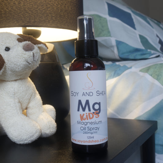 An amber bottle with an atomiser lid stands on a dark bedstand next to a soft plush toy shaped like a dog. A bedside lamp emits a soft glow and bedcovers can be seen in the background.