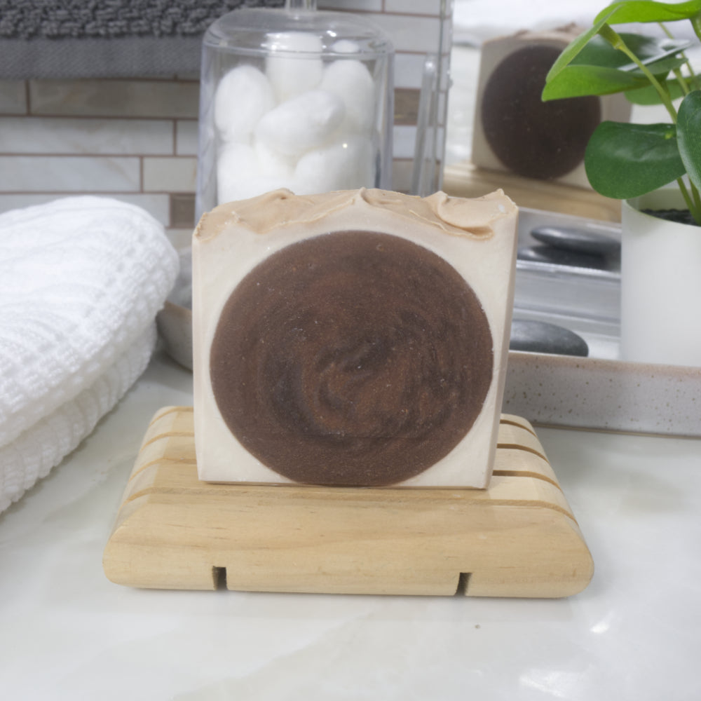 A rectangular bar of soap with a dark brown circular swirl in the center and light beige edges, displayed on a small wooden soap dish on a bathroom counter. A white towel, glass jar, and potted plant are visible in the background.