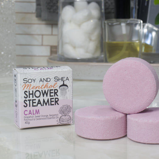 A box of Soy and Shea Menthol Shower Steamer labeled 'Calm' sits on a countertop next to three round, pale mauve shower steamer discs.
