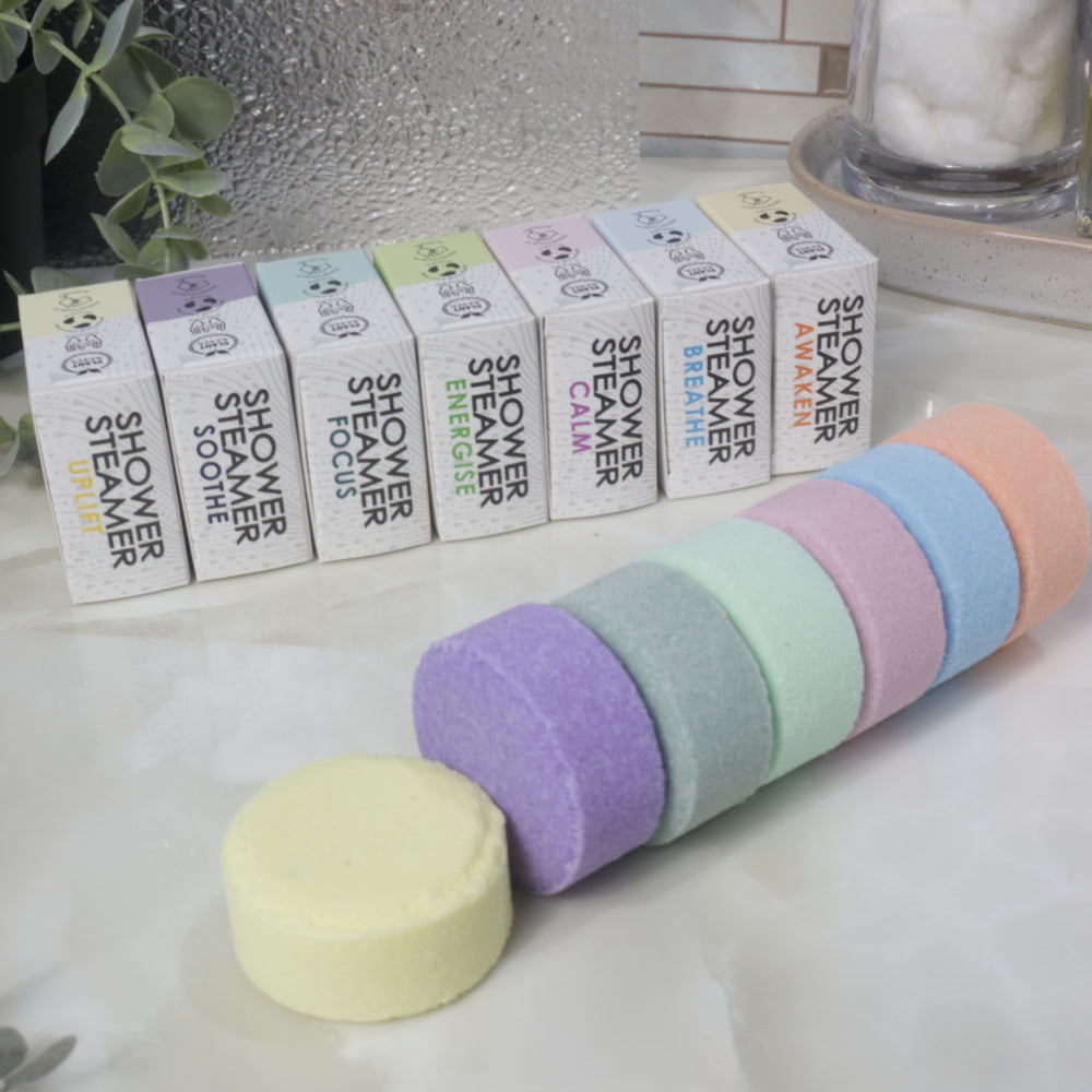 A set of pastel shower steamers, including lavender, mint, light blue, pink, peach, and pale yellow, shown with their matching white packaging boxes on a marble countertop.