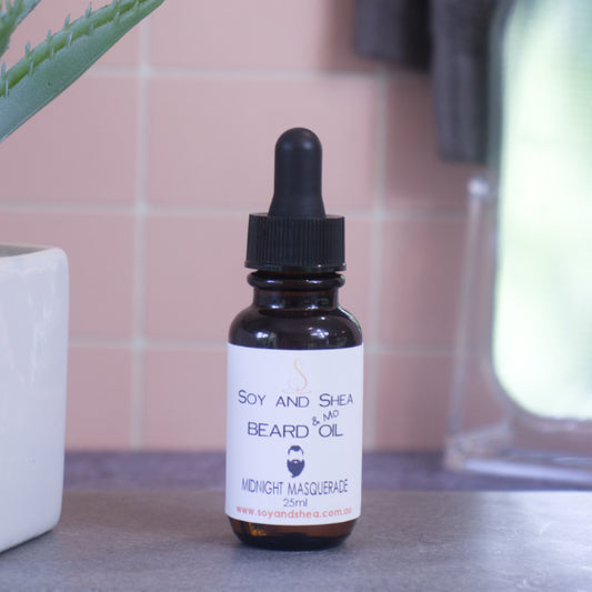 A brown bottle with white label and black dropper lid stands on stone surface next to a pot with green leaves against a blurred tiled background. The label reads BEARD AND MO OIL and shows the image of a man with bead and moustache.