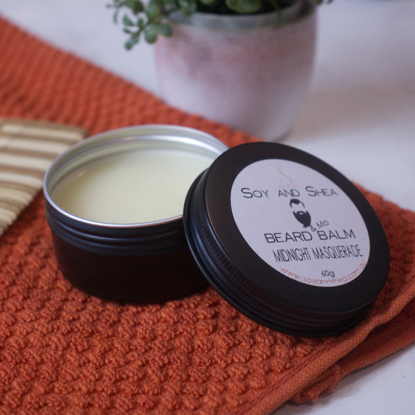 A black tin filled with cream balm sits on an orange towel next to a comb. The black lid rests on the side of the tin with a white round label reading BEARD AND MO BALM
