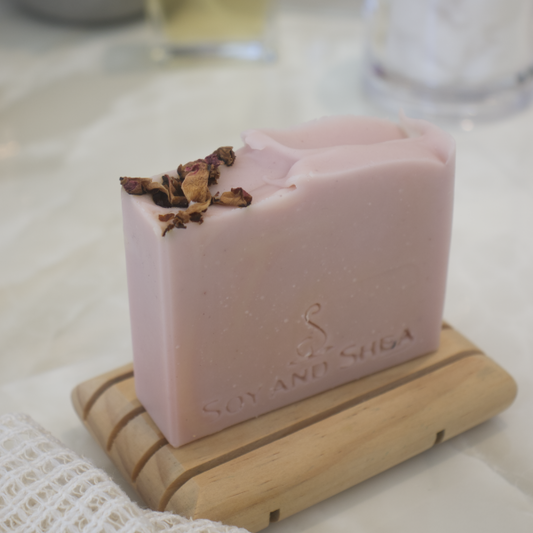 A Dusky Pink Soap bar stamped with SOY AND SHEA featuring some dried rose petals on the top left of the soap sits on a wooden soap dish with a blurred background