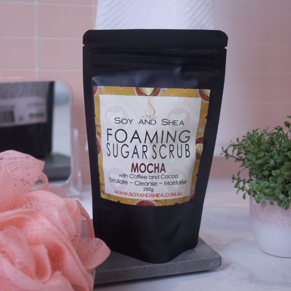 A black zip lock pouch stands upright on a stone tray in front of blurred bathroom background with peach tiles. The pouch, which stands between a potted plant and a loofah, has a label that reads FOAMING SUGAR SCRUB MOCHA and features a ochre background with the top view of filled coffee cups