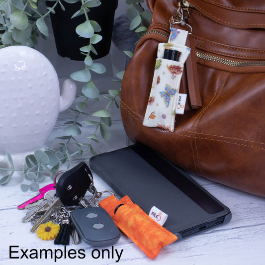 A brown bag sits on a bench along with a set of keys and a mobile phone. Attached to the keys is an orange lip balm holder with a lip balm inside. There is a second holder attached to the bag