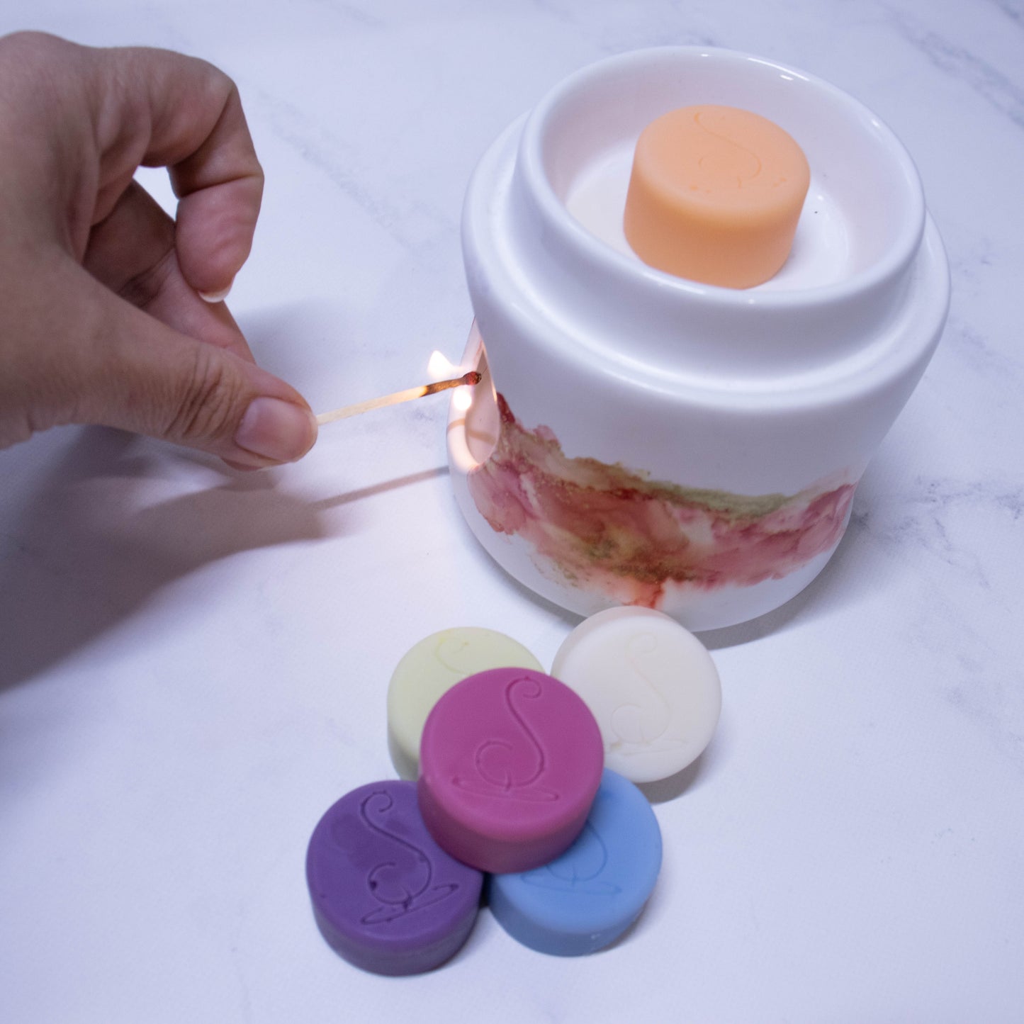 A hand reaches in with a lit match to light an oil burner. In the top sits an orange disc of wax and beside the burner is a stack of various other melts.