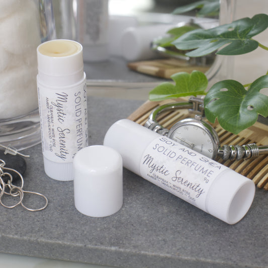 Two white tubes of Solid Perfume are displayed on a grey stone tray along with silver earrings, watch and greenery. One tube lays flat displaying the fragrance notes while another tube stands upright to the left it with the lid removed and to the front revealing a beige balm