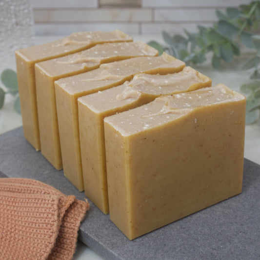 Five bars of soap lined up on a gray surface with a brown textured towel and greenery in the background.