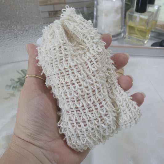 A Hand is holding a rough looking cotton bag against a blurred backdrop of a bathroom counter. The bag is cream with a woven cotton like appearance with little loops on the bag