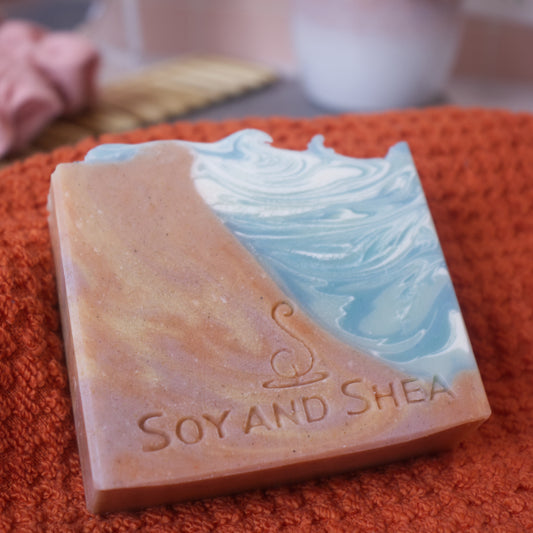 A bar of soap rests on an orange cloth with a blurred background. The left side of the soap has a swirling pattern of brown and gold and to the left is a water like swirl in blues and whites.