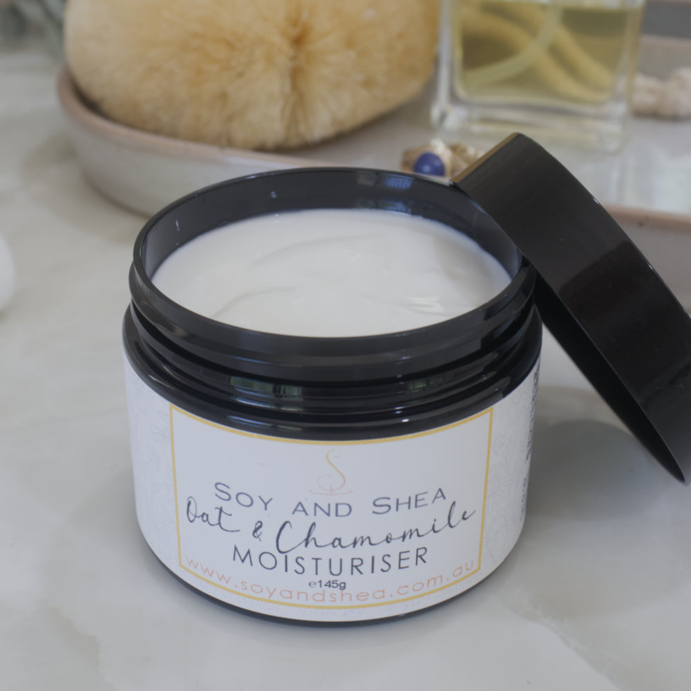 A jar of Oat and Chamomile Moisturiser sits on mable benchtop with a blurred bathroom background. The black lid has been removed from the jar and is resting against the side of the jar revealing a white cream.