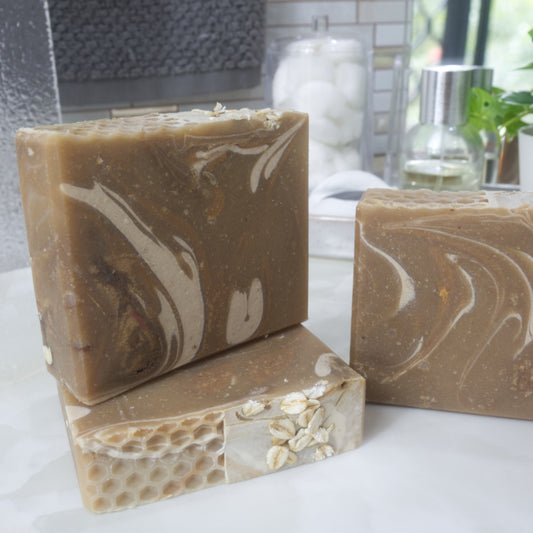A collection of soaps are presented on a bathroom counter with blurred background. One soap lays flat on the bench showing the soap top with a honeycomb pattern and oat pieces. On top stands another soap and to the right is a glimpse of a third soap. All soaps are a warm gold colour with swirls of Cream and rich gold