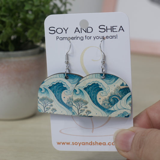 A hand holding a branded Soy and Shea card featuring a pair of earrings. The earrings are a solid arch shape with a blue wave design.