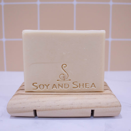 A bar of soap stands on a wooden soap dish on a white surface with peach tile background. The soap is a solid cream colour and stamped with logo and SOY AND SHEA