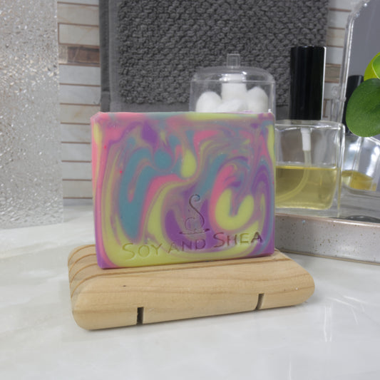 A rectangular bar of soap stands on a wooden soap rack on a bathroom countertop next to a bottle of perfume and jar of cotton balls. The soap has a rainbow swirl of bright neon colours in Yellow, blue, pink and purple