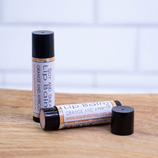 Two black tubes of lip balm are displayed on a wooden surface with blurred tiled background. One tube stands on its end while the other lays flat in front with the cap removed revealing a tan balm. The label is pale orangewith illustrations of orange and apricots