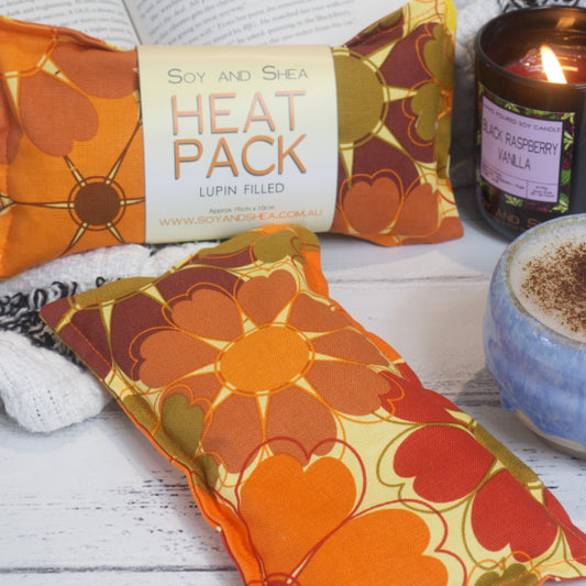 Two Orange Bloom Mini Heat Packs by Soy and Shea rests near a ceramic cup, a burning candle, and an open book—creating an inviting cosy scene. The Mini Heat packs feature a bright orange and red flowers with heart shaped petals scattered on a pale yellow background