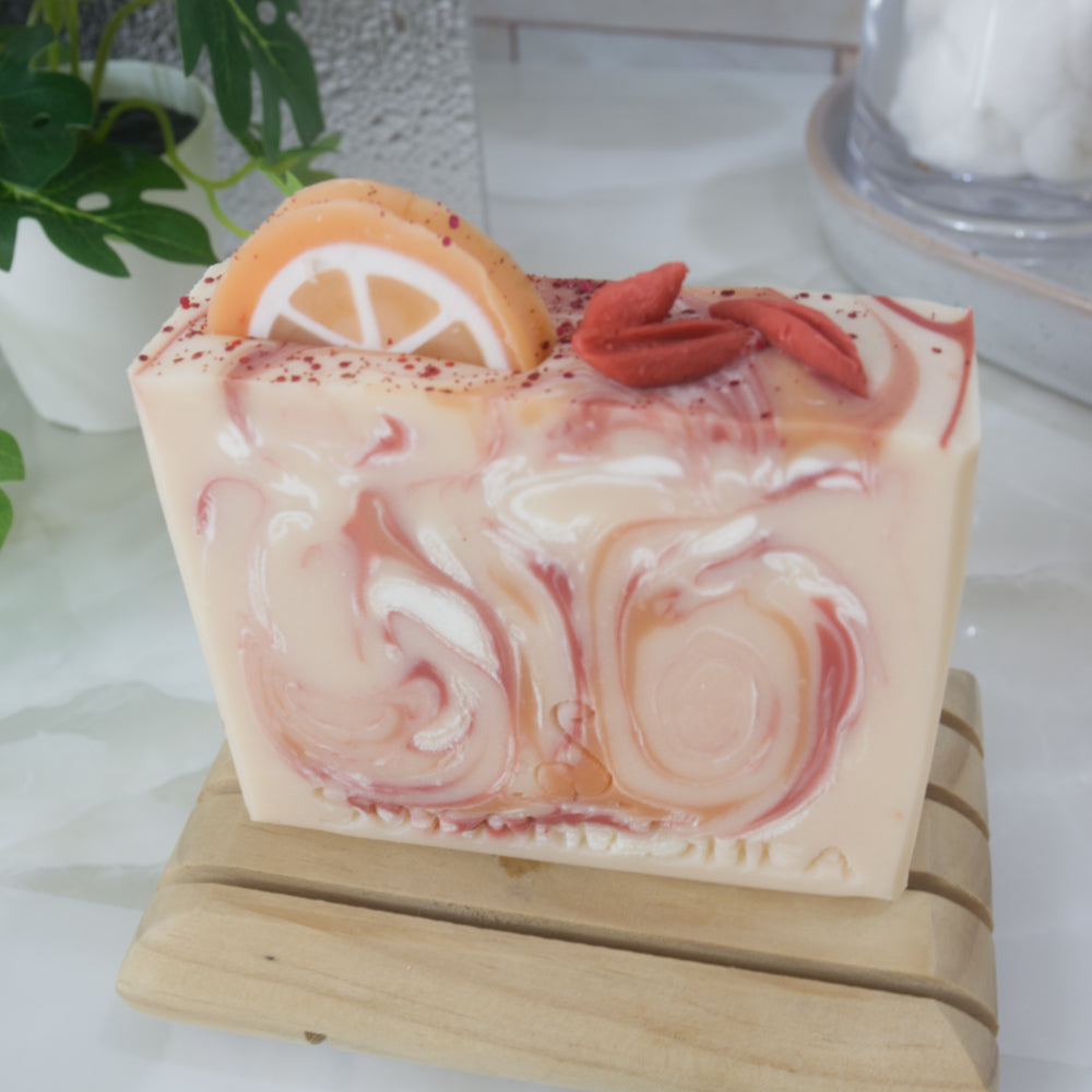 An soap bar stands on a wooden soap dish on a bathroom countertop with a plant and stone tray. The soap is pale orange with swirls of bright orange, red and white. The image focuses on the top of the soap which features soap decorations of an orange slice and some red berries and some red biodegradable glitter
