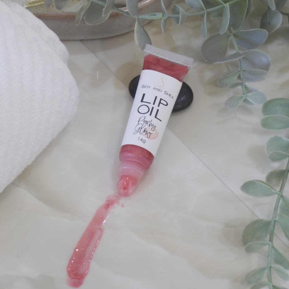 A tube of Lip Oil lies on a marble tile surface next to a white towel and some eucalyptus leaves. The tube is clear with a white product label and filled with a peachy red Lip oil. The cap is removed and a smear of Lip oil is on the bench showing the translucent effect it has