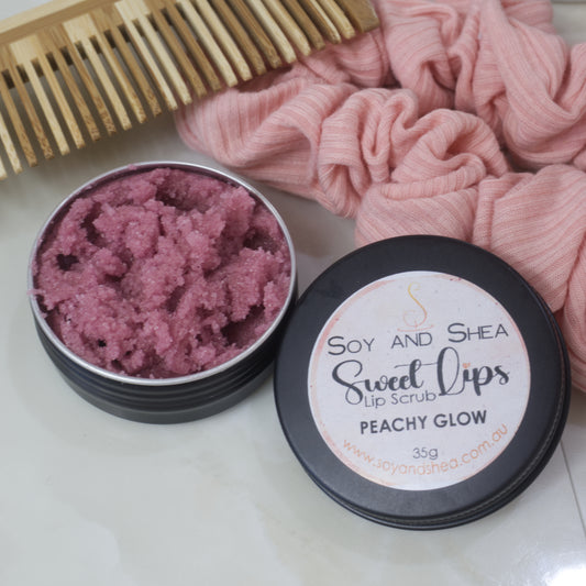 A black tin of Lip Scrub sits on a marble countertop next to a scrunchie and wooden comb. The lid has been removed and rests on the side of the tin revealing a dusky pink scrub