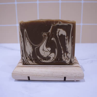A bar soap stands on a wooden soap dish on white surface with a faded peach tile background. The Soap has a rich brown base colour with cream swirls