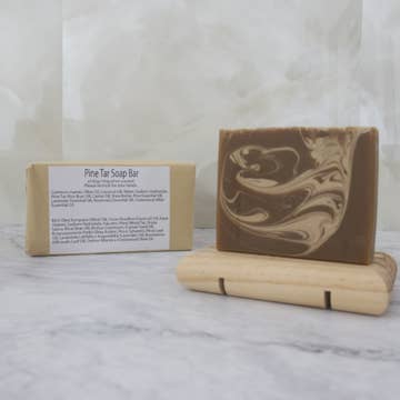 A brown soap bar with light brown swirls sits on a wooden stand with a brown paper package behind labeled 'Pine Tar Soap Bar' on a marble background