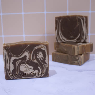 A collection of soaps sit on a white marble surface against a pale peach tile background. In the background, two soaps lay flat but stacked upon each other revealing a flat textured top. A third soap stands on stop of this stack with a fourth soap in the foreground. The soaps are a rich brown colour with unique cream swirls