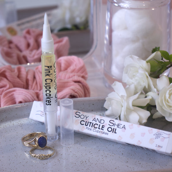 A clear, narrow tube with the lid removed showing a brush tip stands upright on a stone tray. Beside it sits a cardboard box labelled CUTICLE OIL and a set of gold rings. In the background is a bathroom counter top with a pink scrunchie, small mirror, jar of cotton balls and a some white flowers.