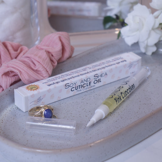 A clear, narrow tube with the lid removed showing a brush tip lies on a stone tray beside a cardboard box labelled CUTICLE OIL and a set of gold rings. In the background is a bathroom counter top with a pink scrunchie, small mirror, and a some white flowers.
