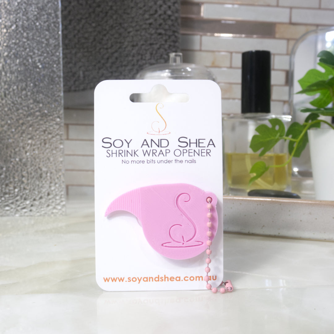 A light pink Shrinkwrap opener with matching ball and chain keyring is attached to a white card with product details. The card is standing upright on a bathroom countertop with a bottle of perfume, a jar of cotton balls and towel in the background