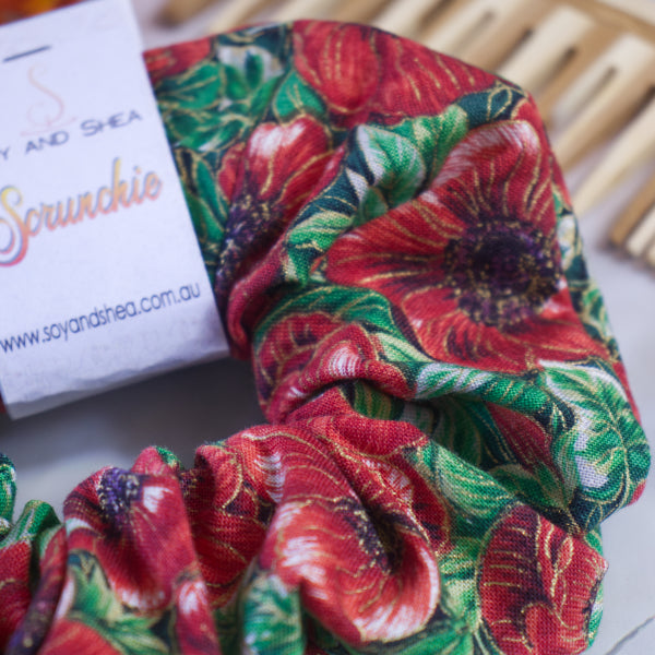 This vibrant, handmade scrunchie by Soy and Shea showcases a lively red and green floral design. A partially visible label reads "Sprunchie," complete with a website ending in ".com.au." Its unique knotless elastic construction is set against a background that suggests the texture of wicker.