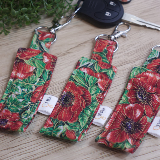 A selection of three lip balms showing variations in fabric design, lie on a wooden side with one attached to a car key with a lobster claw clasp. The Clip balms have a bold pattern of large red poppies with purple/black centres surrounded by bright green leaves.