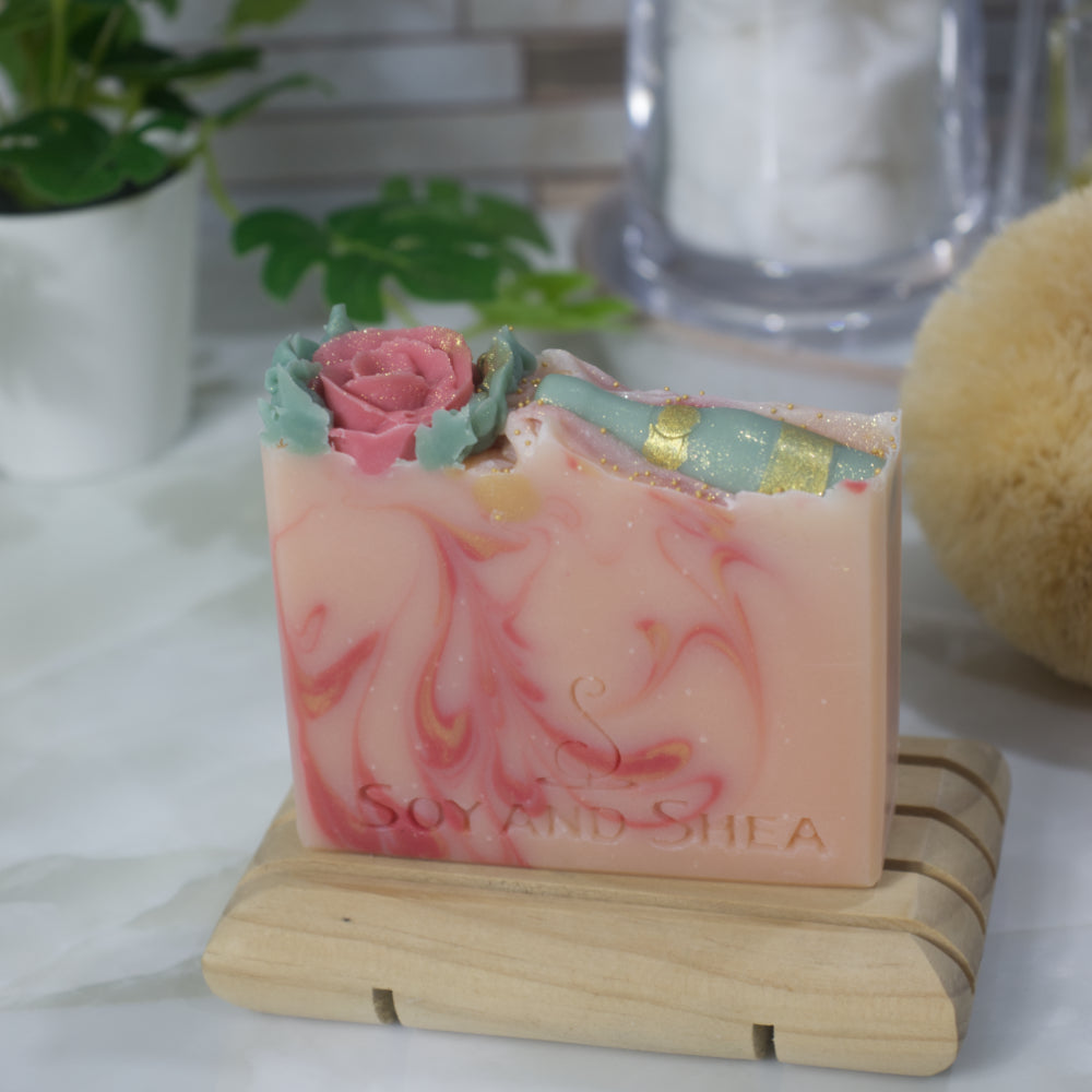 A soap bar stands on a wooden soap rack surrounded by a plant, a jar of cotton balls and body scrubber. The soap is pale pink with dark pink and gold swirls. The top of the bar has soap decorations of single red rose on once side and a green wine bottle with gold accents on the other.