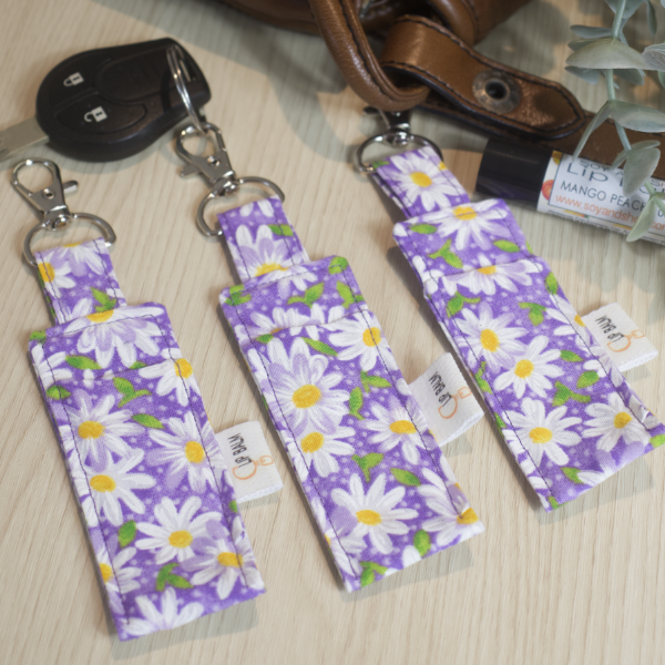 A selection of three clip balms showing variations in fabric design, lie on a wooden side with the middle Clip Balm attached to a car key. A lip balm lays near-by along with some greenery. The Clip balms are purple with a scattering of white daisies with yellow centers and green leaves. Each